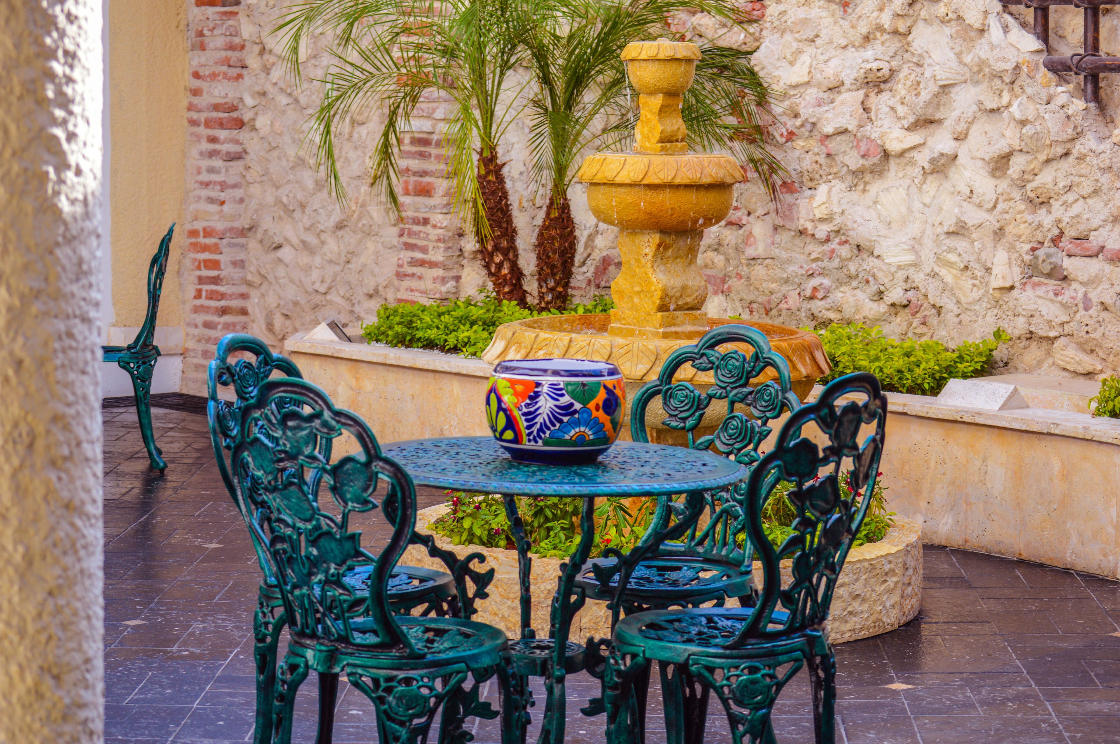 A courtyard with a round table and four chairs, a decorative fountain, and a brick wall with plants.