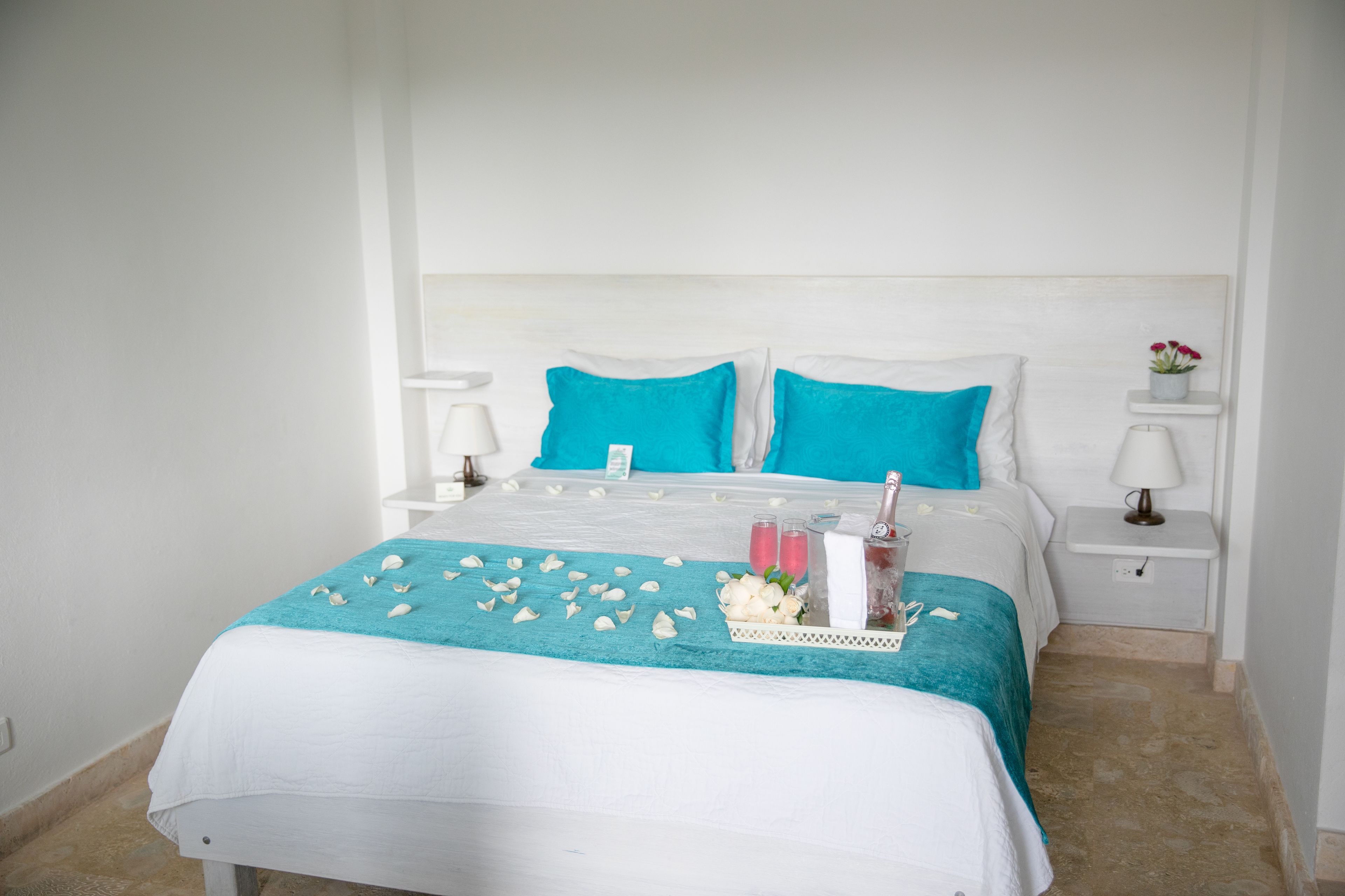A neatly made bed with a white and teal bedspread, two teal pillows, and a bedside table with a lamp and a small vase with flowers.