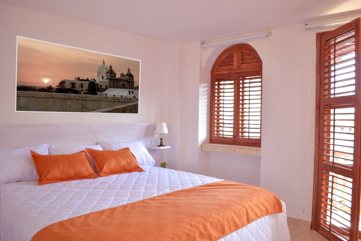 Une chambre d’hôtel avec un lit, une fenêtre avec des volets en bois et un tableau représentant un bâtiment accroché au mur.