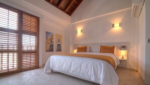 A bedroom with a large bed, wooden shutters, two wall-mounted lights, and a nightstand with a lamp. - Hotel Boutique La Artillería (Cartagena)