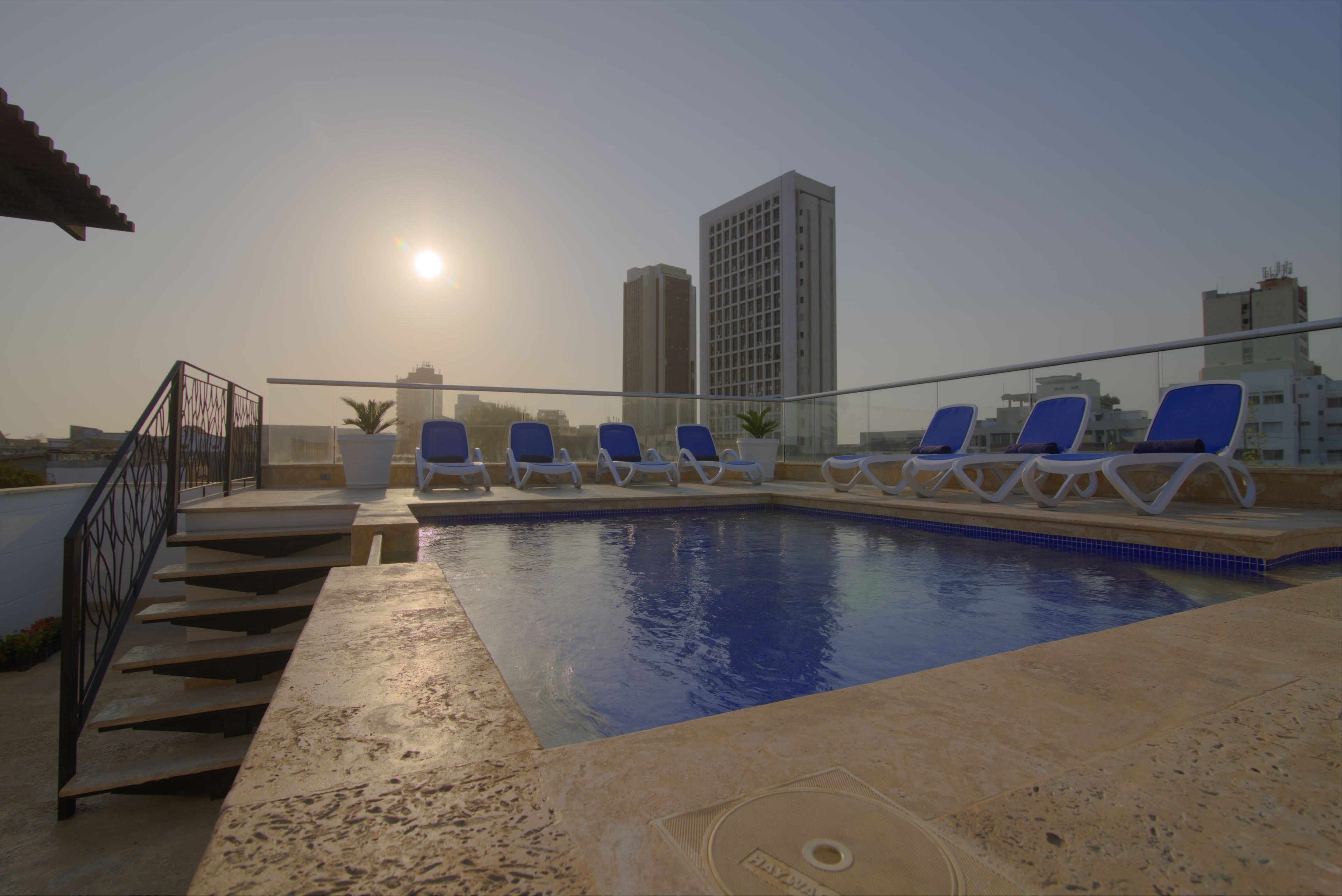 A rooftop pool with blue lounge chairs, a staircase, and a tall building in the background.