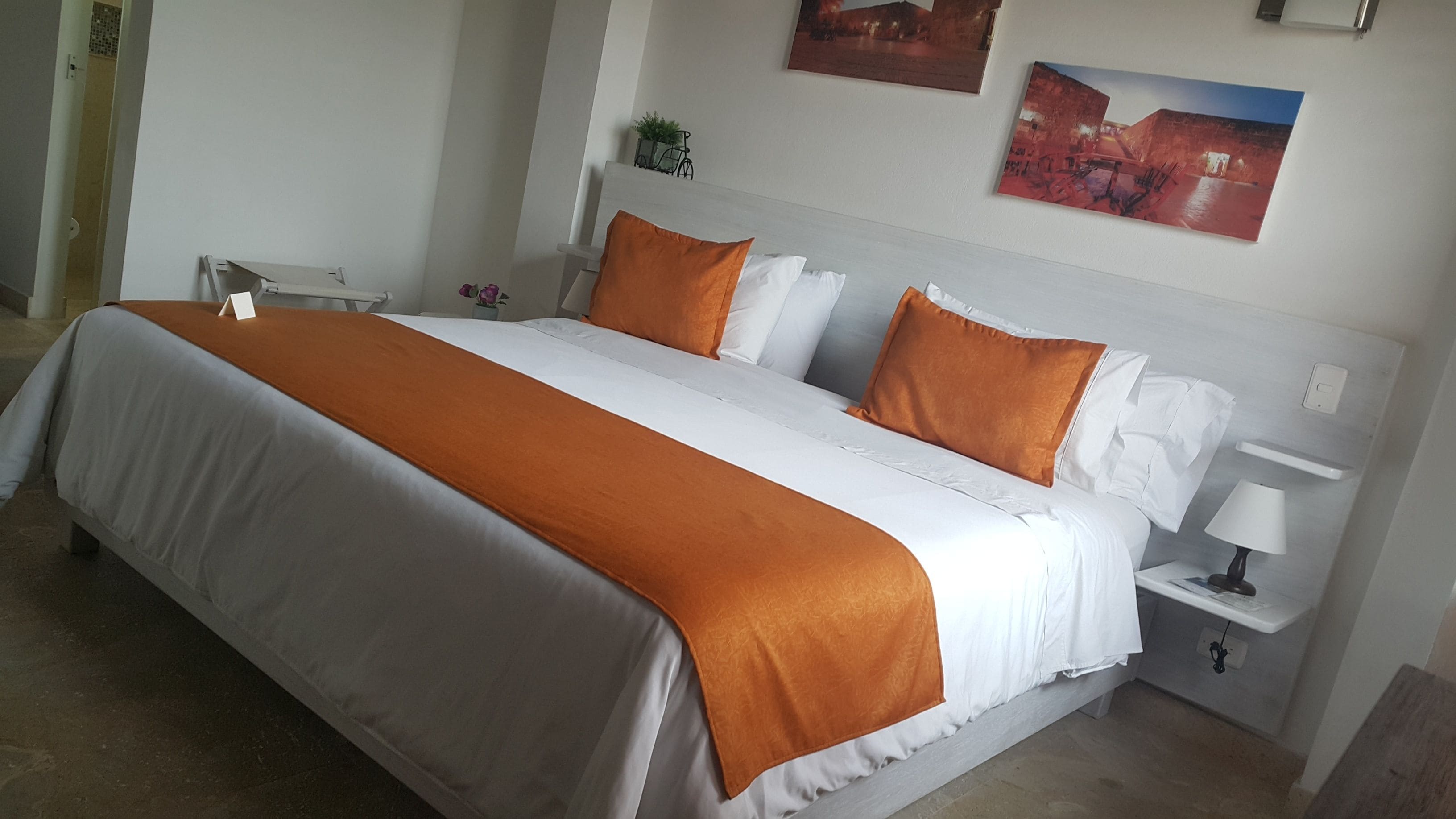 Un lit bien fait, recouvert d’une couvre-lit blanche et orange, avec une tête de lit blanche et deux tables de chevet blanches, chacune surmontée d’une lampe.