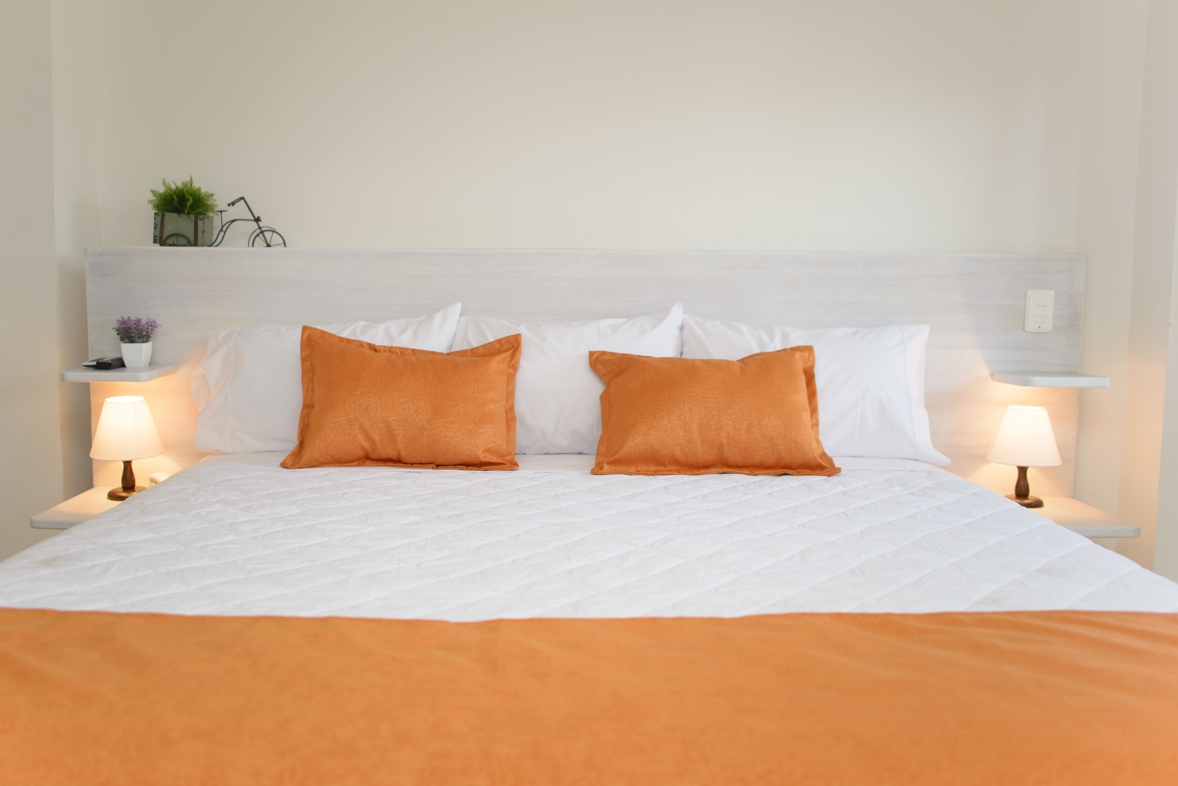 A neatly made bed with white linens and two orange pillows, flanked by bedside tables with lamps and small decorative items.