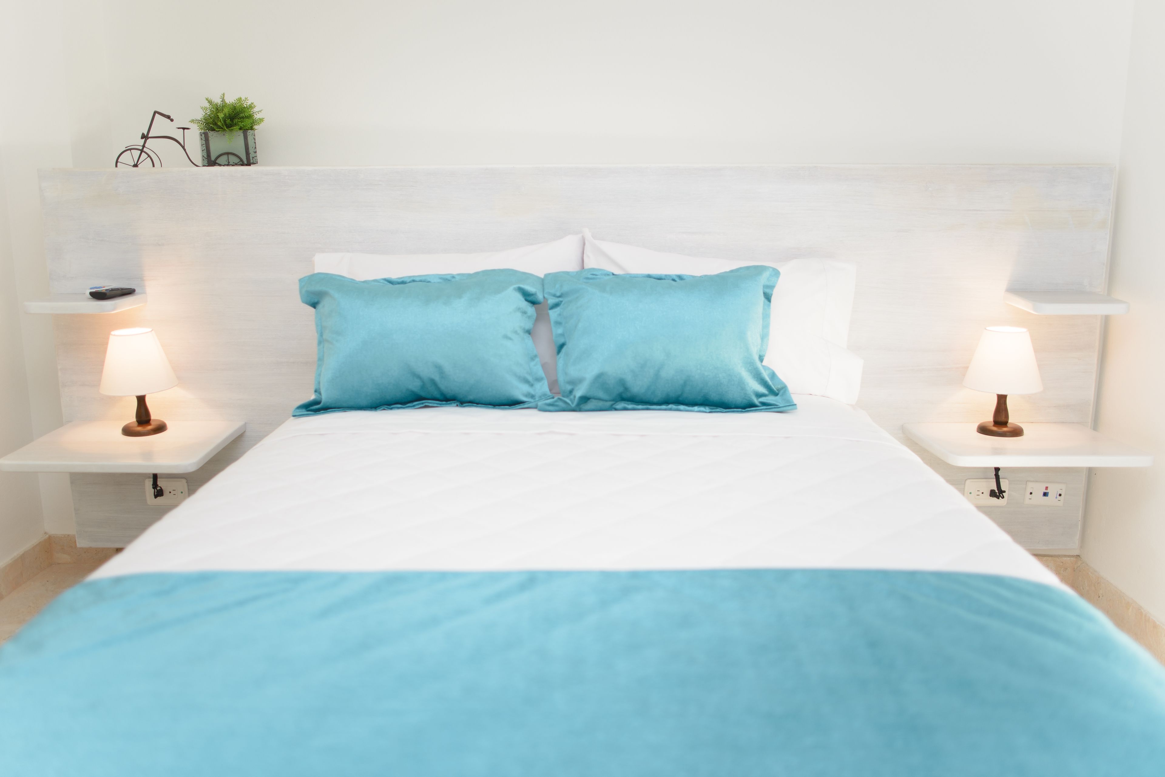 A neatly made bed with two pillows, two bedside tables with lamps, a small shelf with a bicycle and a plant, and a white headboard.