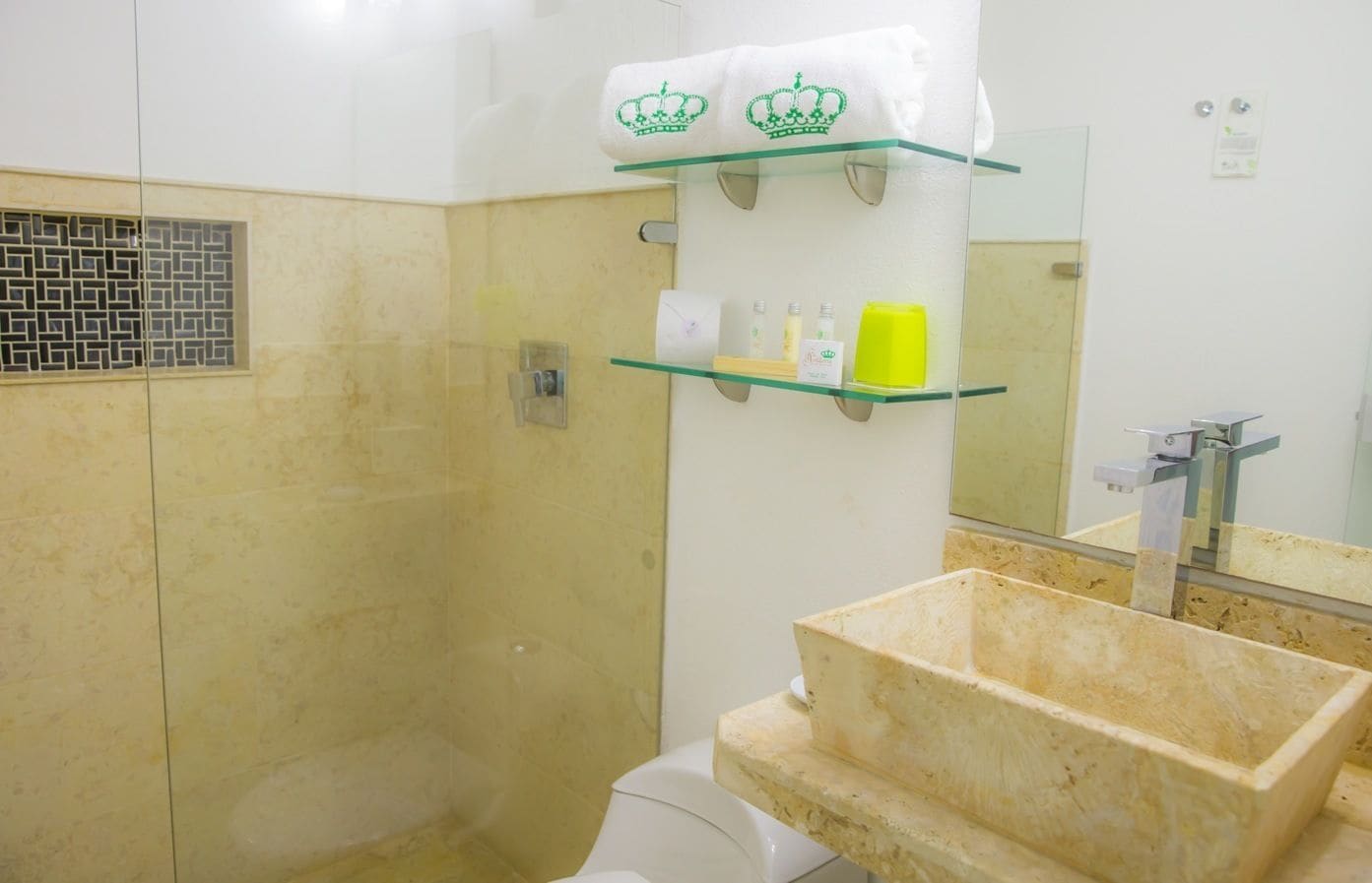 Une salle de bain avec une douche carrelée de couleur beige, une étagère en verre où sont rangés les articles de toilette, un lavabo avec un miroir et des toilettes.