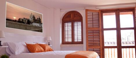 A bedroom with a large bed, a nightstand with a lamp, a window with wooden shutters, and a wall-mounted picture of a building.