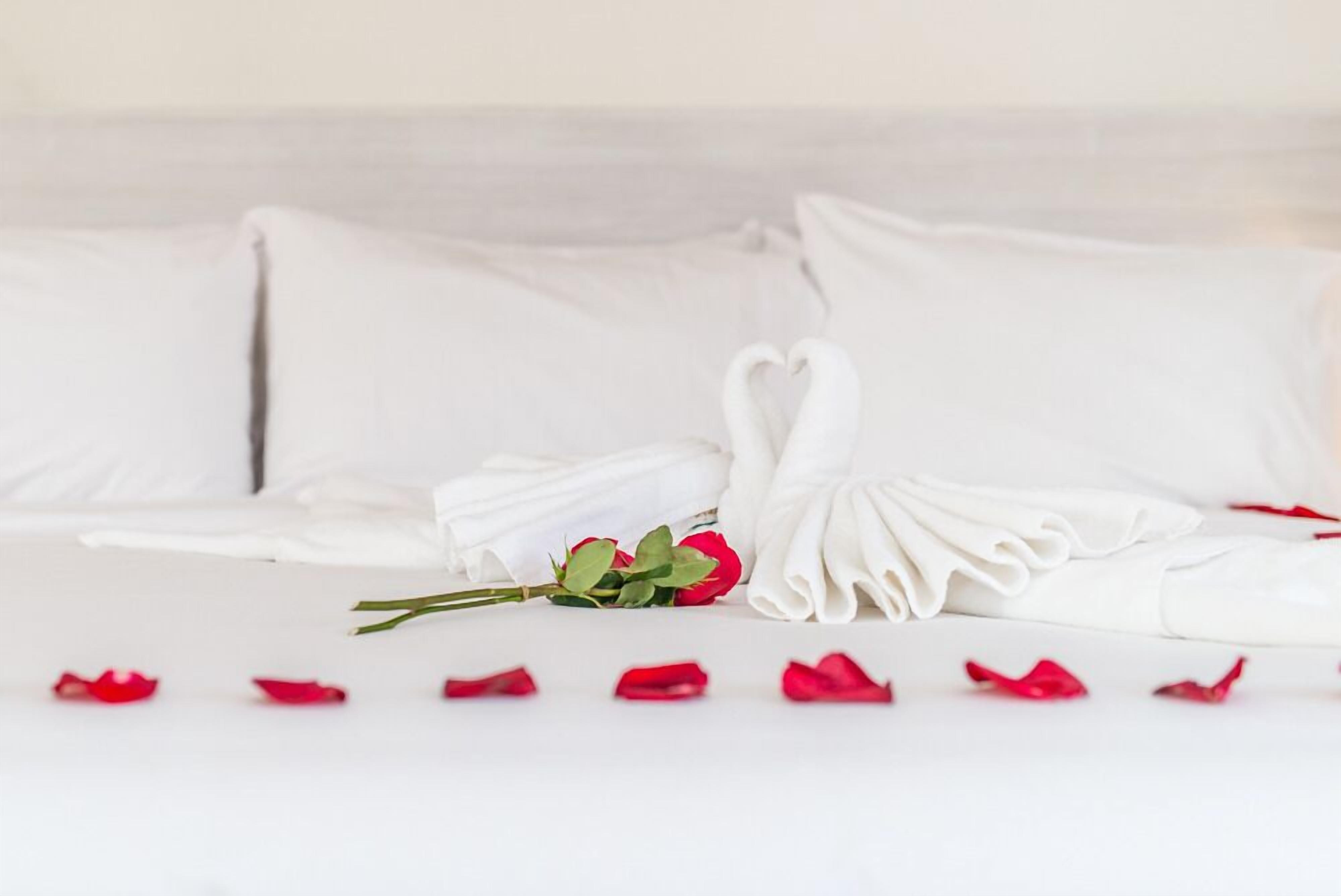 A neatly made bed with white linens, a heart-shaped towel arrangement, and a bouquet of red roses.