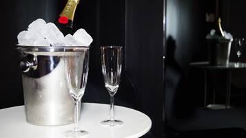 Una bottiglia di champagne in un secchio di metallo con cubetti di ghiaccio, un bicchiere trasparente accanto e un tavolo bianco su sfondo scuro.