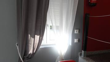 Een kamer met raam, gordijnen, een poster van een rode telefooncel, een brandblusser en een rode stoel.