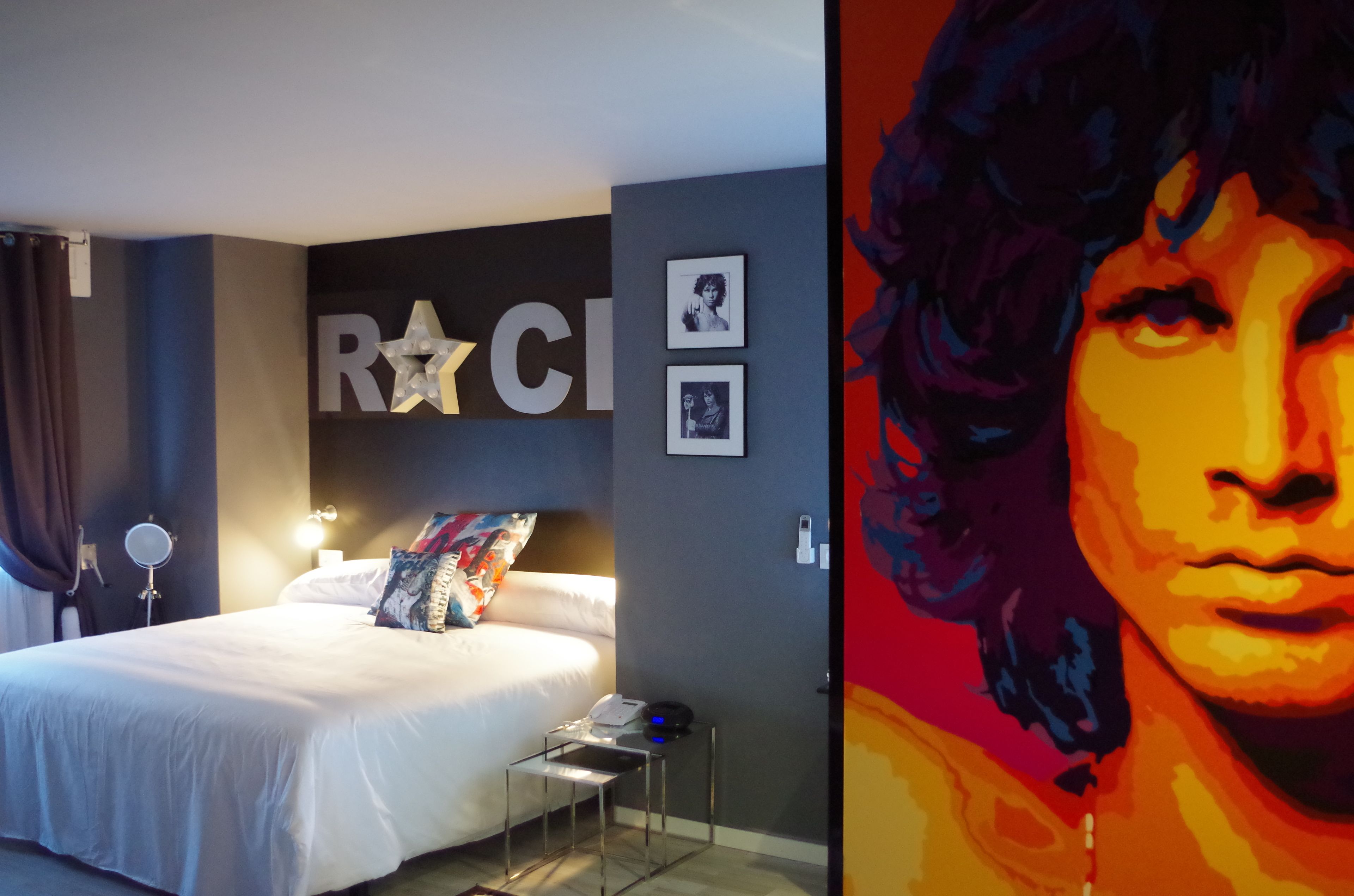 Quarto de hotel moderno com uma cama, uma mesinha de cabeceira, um espelho e uma parede com uma grande estrela, onde se lê parcialmente a palavra 'RAC'.
