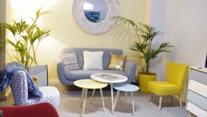 Una sala de estar con un sofá, dos mesas auxiliares, un sillón amarillo y dos plantas en macetas.