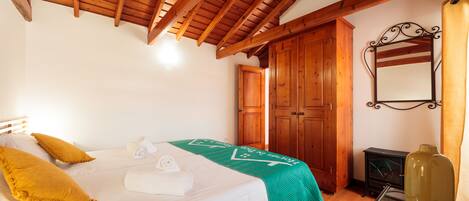 A bedroom with a wooden ceiling, a bed with white bedding and yellow pillows, a green throw blanket, a wooden wardrobe, a mirror, and a green vase on a table.