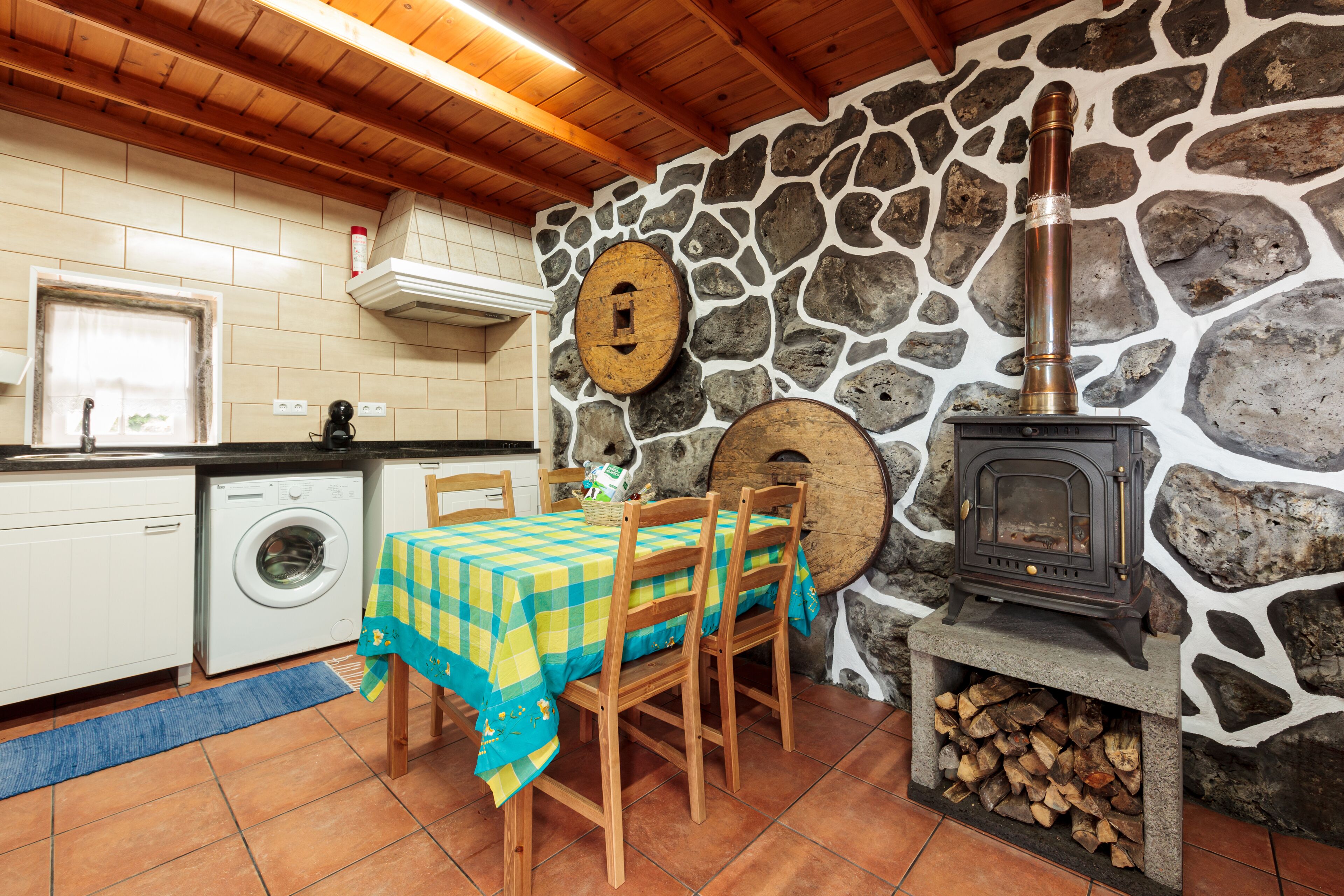 A kitchen with a stone wall, a washing machine, a table with chairs, and a wood-burning stove.