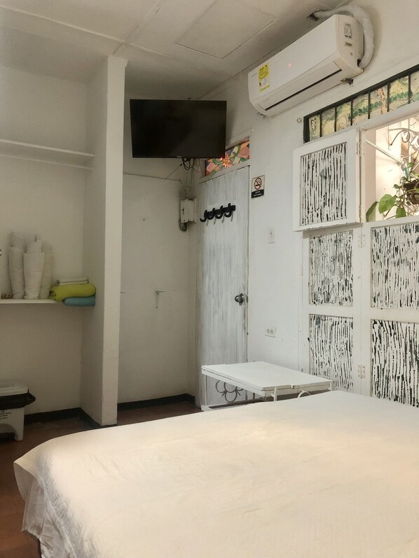 A bedroom with a bed, a small table, a shelf with folded clothes, an air conditioning unit, and a door with a 'No Smoking' sign.