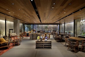 A spacious interior with wooden ceiling and large windows, featuring a central coffee table with books and a lamp, surrounded by wooden chairs and tables.