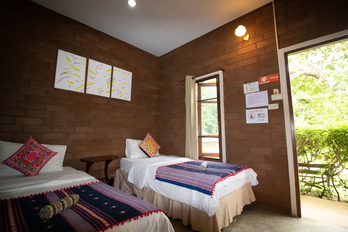 A room with two beds, a brick wall, and a window with a view of greenery.