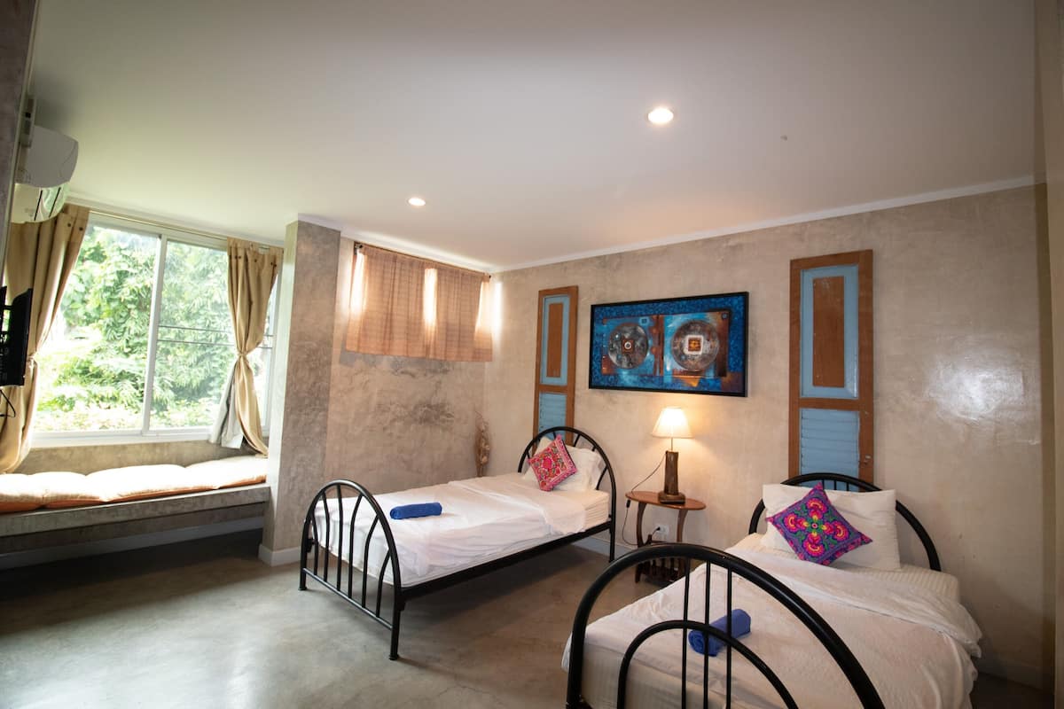 A room with two single beds, a window with curtains, a painting on the wall, and a small table with a lamp.