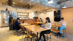 Un café moderno con mesas y sillas de madera, un área de cocina y una pared con una fecha y el nombre de un evento.