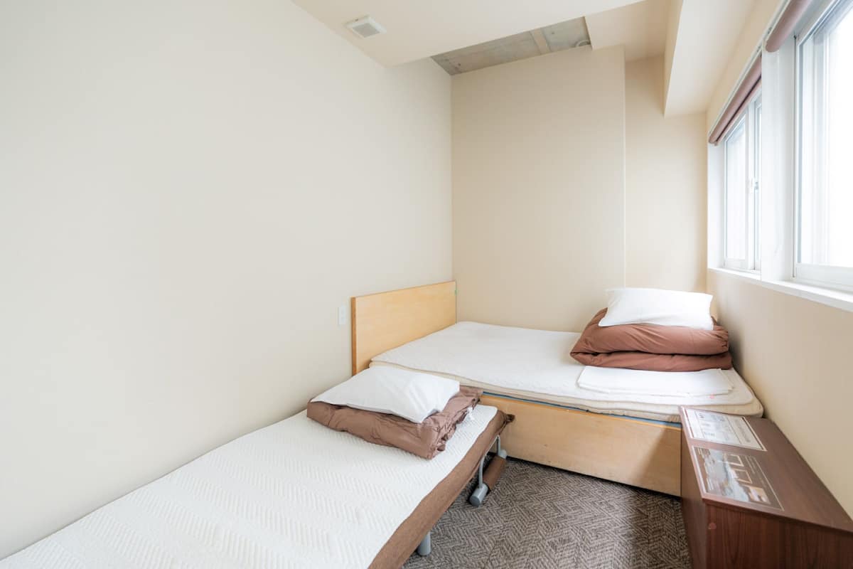 A small, single-bed room with a wooden headboard, a window with blinds, and a small desk with a chair.