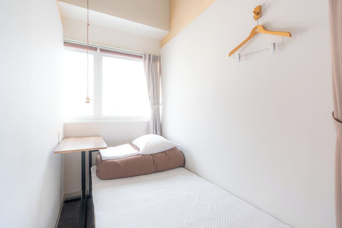 A small, neatly arranged bedroom with a single bed, a bedside table, a hanging light, and a wooden hanger on the wall.