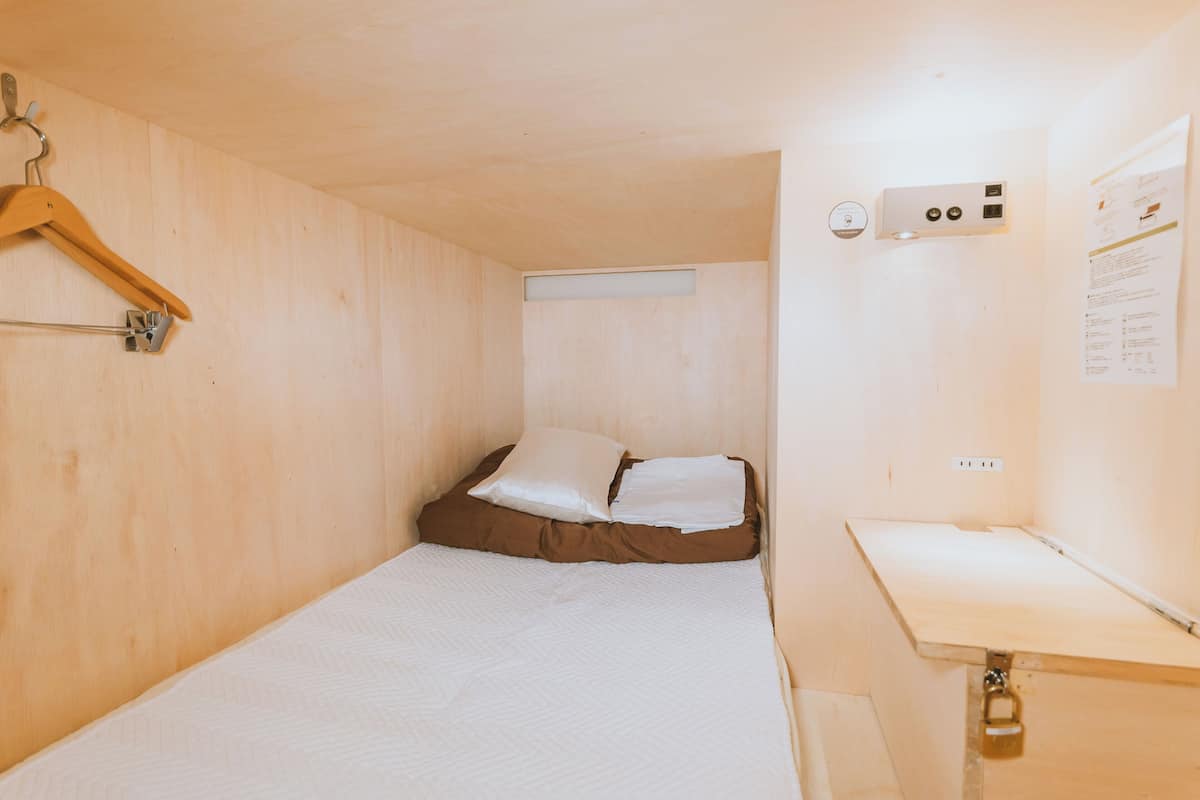 A small, single-bed room with a wooden bunk bed, a wooden hanger, a small table, and a wall-mounted heater.