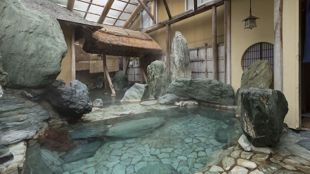 A traditional Japanese onsen with a stone bath surrounded by rocks and a wooden structure with a thatched roof. - Awara Grand Hotel (Awara)