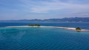A coastal landscape with a sandy beach, a small island with trees, and distant mountains under a clear sky. - Awara Grand Hotel (Awara)