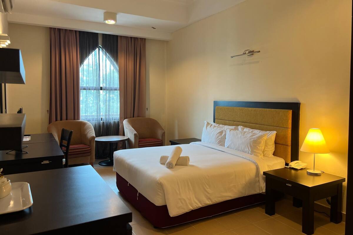 Ein Hotelzimmer mit Bett, Schreibtisch, Stuhl, einem kleinen Tisch, einer Lampe und einem Fenster mit Vorhängen.