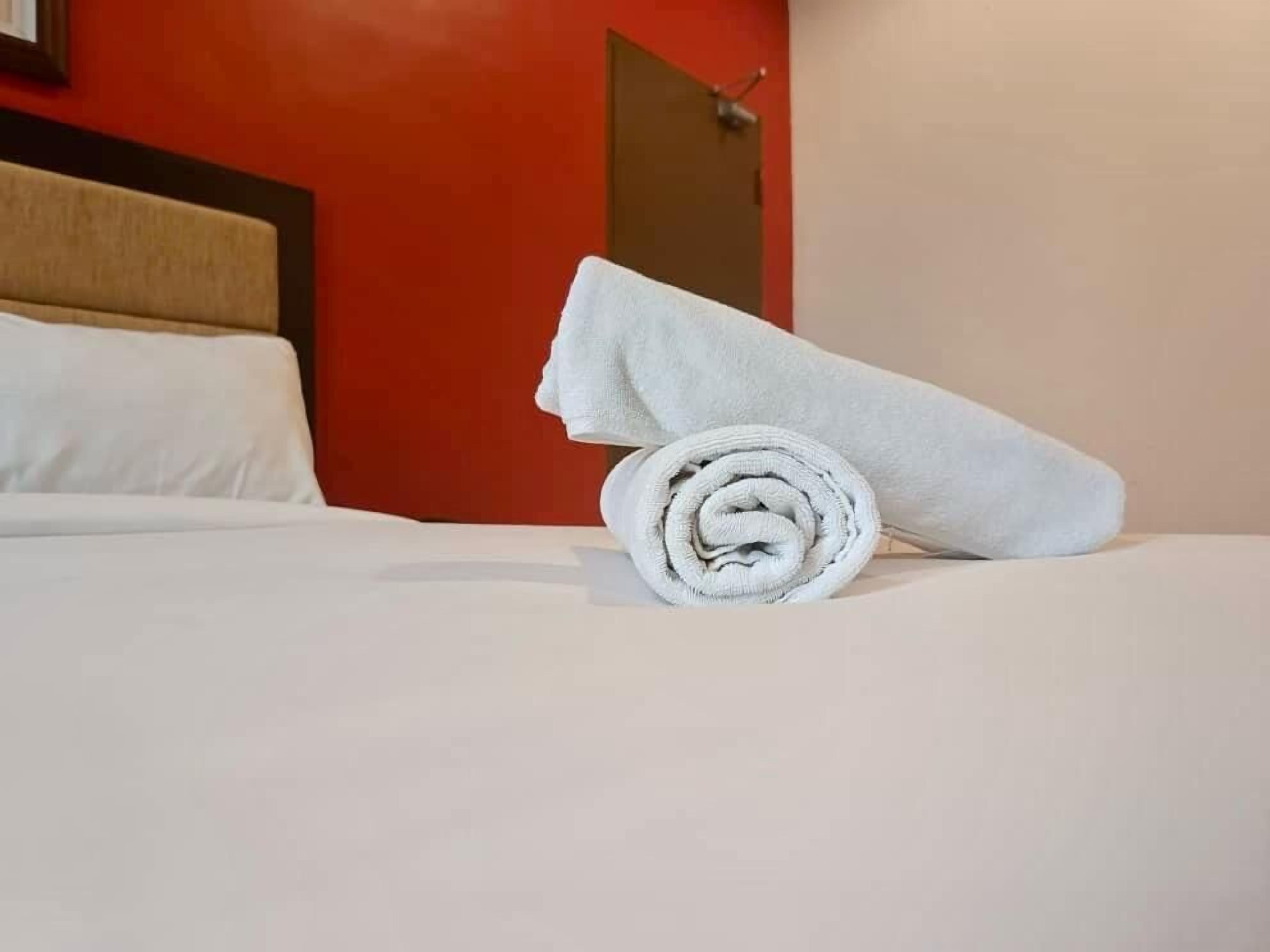 A neatly made bed with a rolled white towel on it, a red wall, and a door in the background.