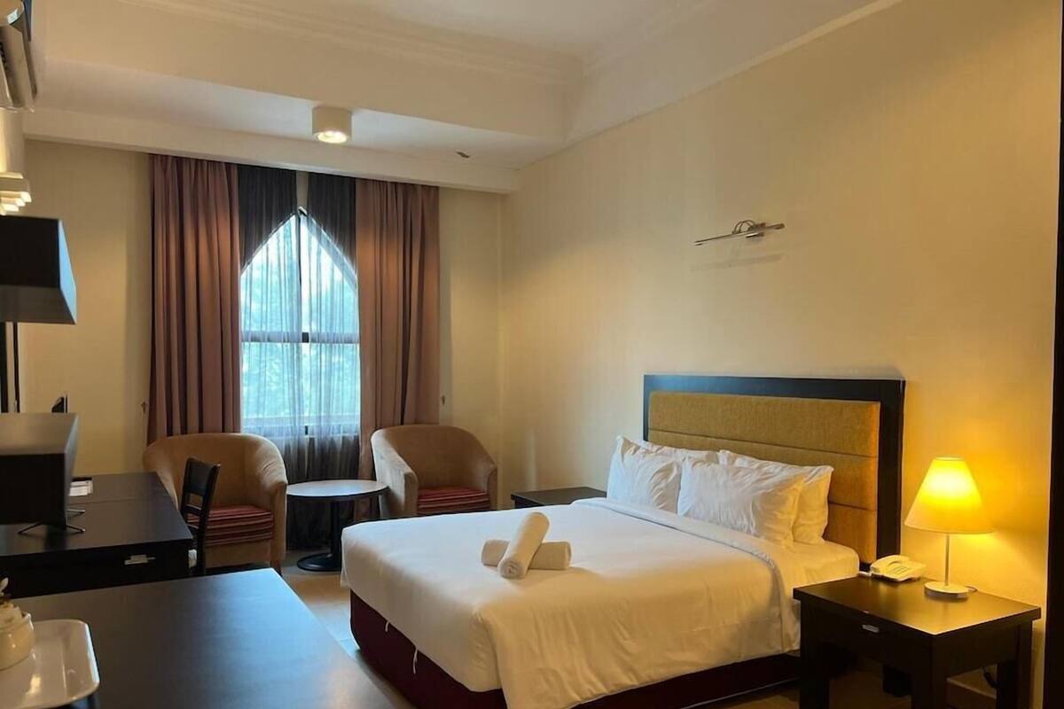 Ein Hotelzimmer mit einem Bett, einem Schreibtisch, einem Stuhl, einer Lampe und einem fenster mit Vorhängen.