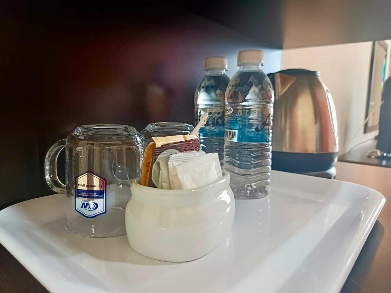 Un plateau avec une tasse en verre, un récipient blanc contenant des sachets et deux bouteilles d’eau.