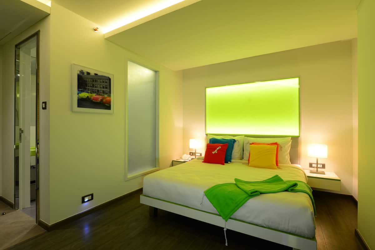 Une chambre d’hôtel avec un grand lit, deux tables de chevet avec des lampes, un lampadaire vert fixé au mur au-dessus du lit et un tableau encadré accroché au mur.