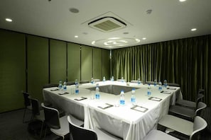 Un salón de conferencias con una mesa blanca, sillas, botellas de agua y un fondo de cortina verde.