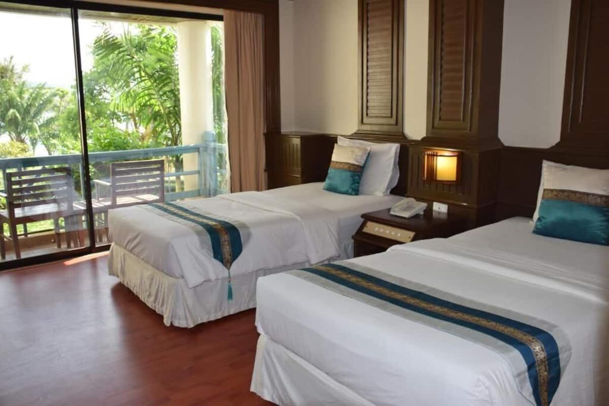 A hotel room with two beds, a balcony with wooden chairs, and a view of palm trees.