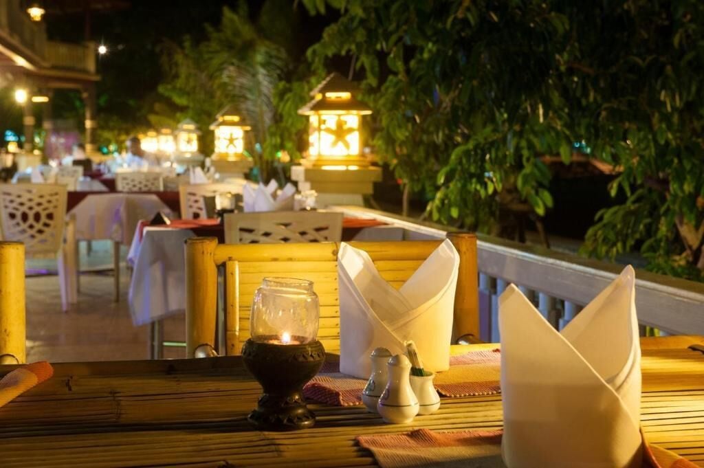 An outdoor dining area with tables set for guests, illuminated by lanterns, and surrounded by greenery.