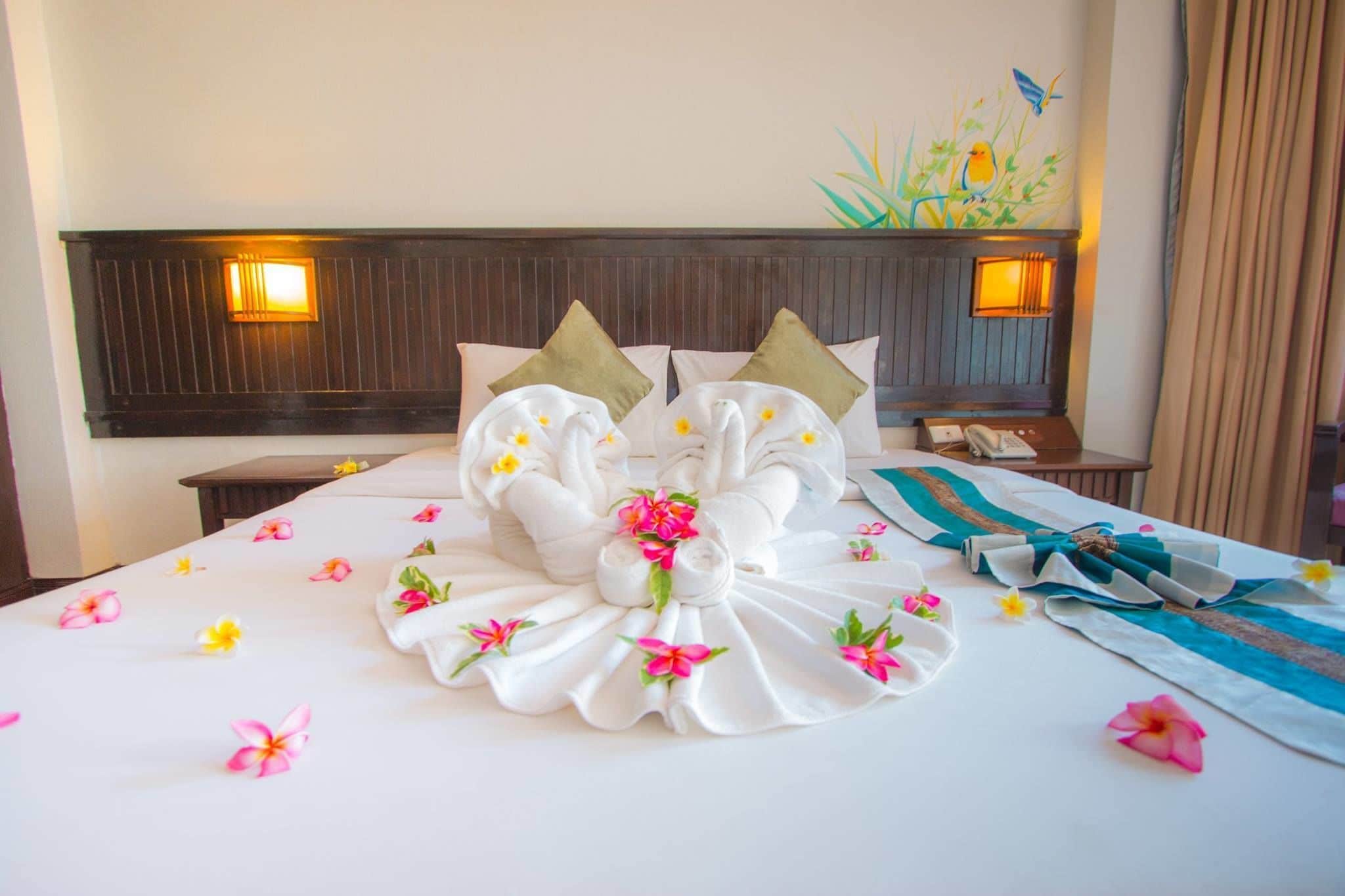A neatly made bed with a floral arrangement, two pillows, a bedside table with a phone, and a wall-mounted light fixture.