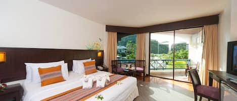 A hotel room with a large bed, a desk with a TV, and a balcony with a view of greenery.