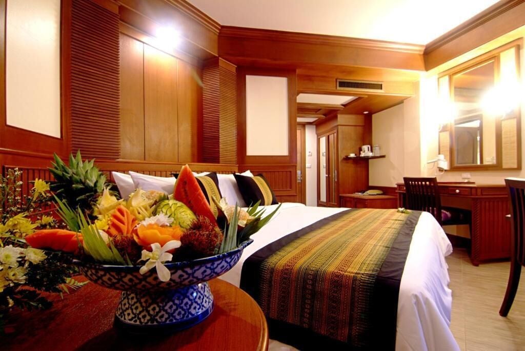 A hotel room with a bed, a desk, and a dining area. There is a bowl of fruit on the bed.