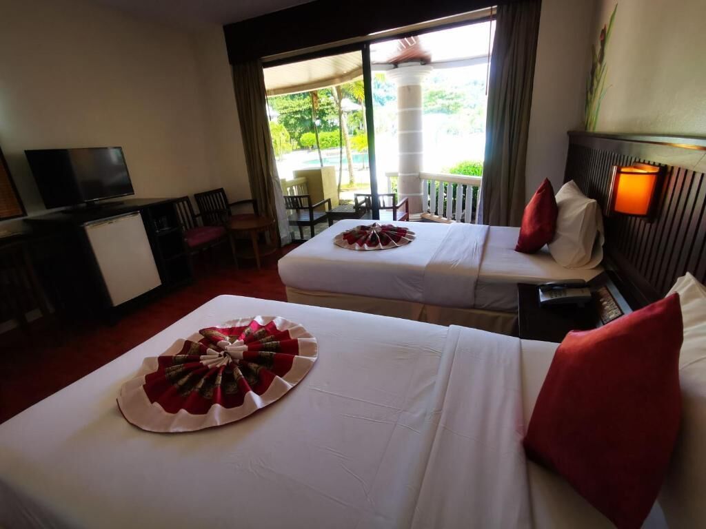 A hotel room with two beds, a TV, a desk, and a balcony with a view of greenery.