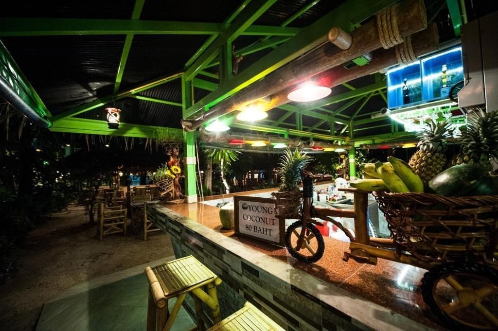 Un restaurant avec un toit en métal vert, une enseigne indiquant « Noix de coco jeune, 80 bahts », et une étalage d’ananas et de bananes.