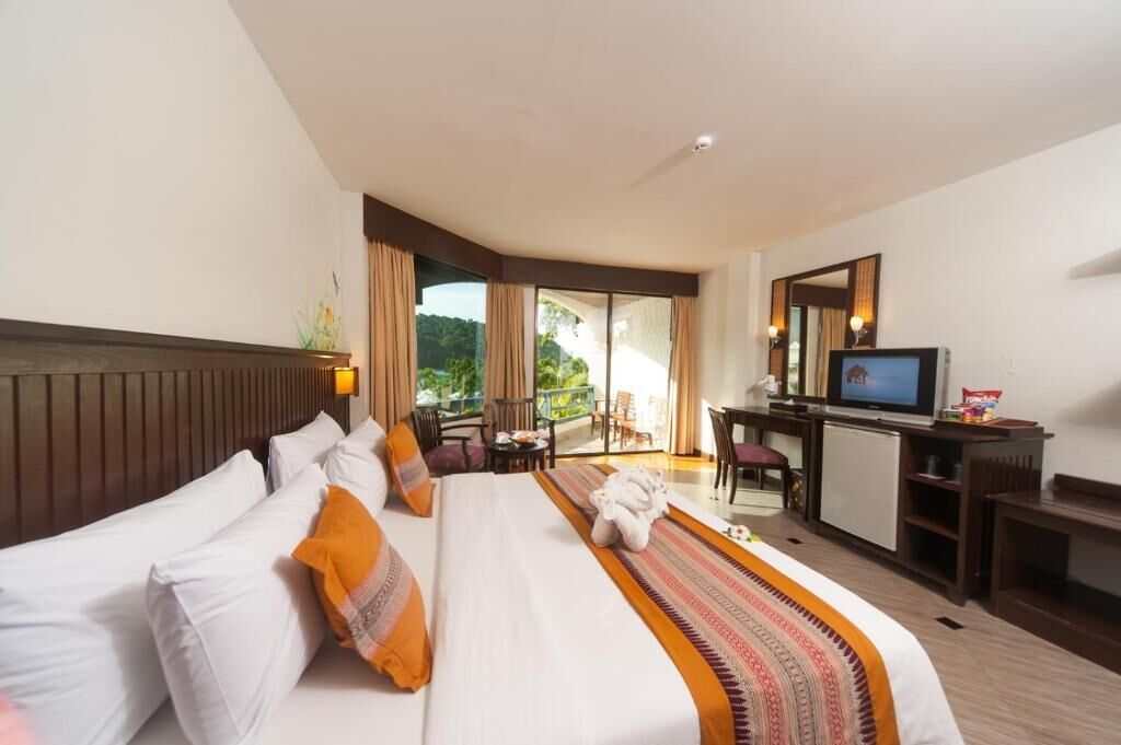 A hotel room with a large bed, a desk, a television, and a balcony with a view of greenery.