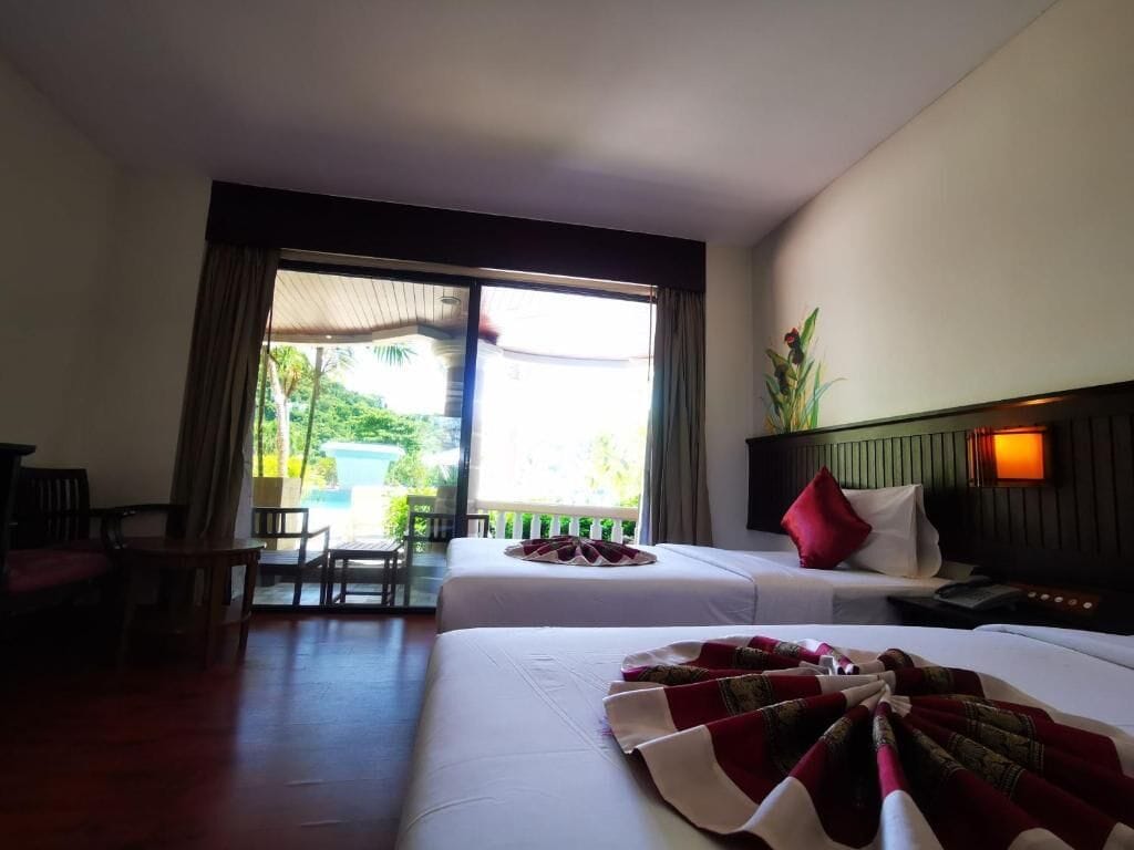 A hotel room with two beds, a dining area, and a view of greenery outside.