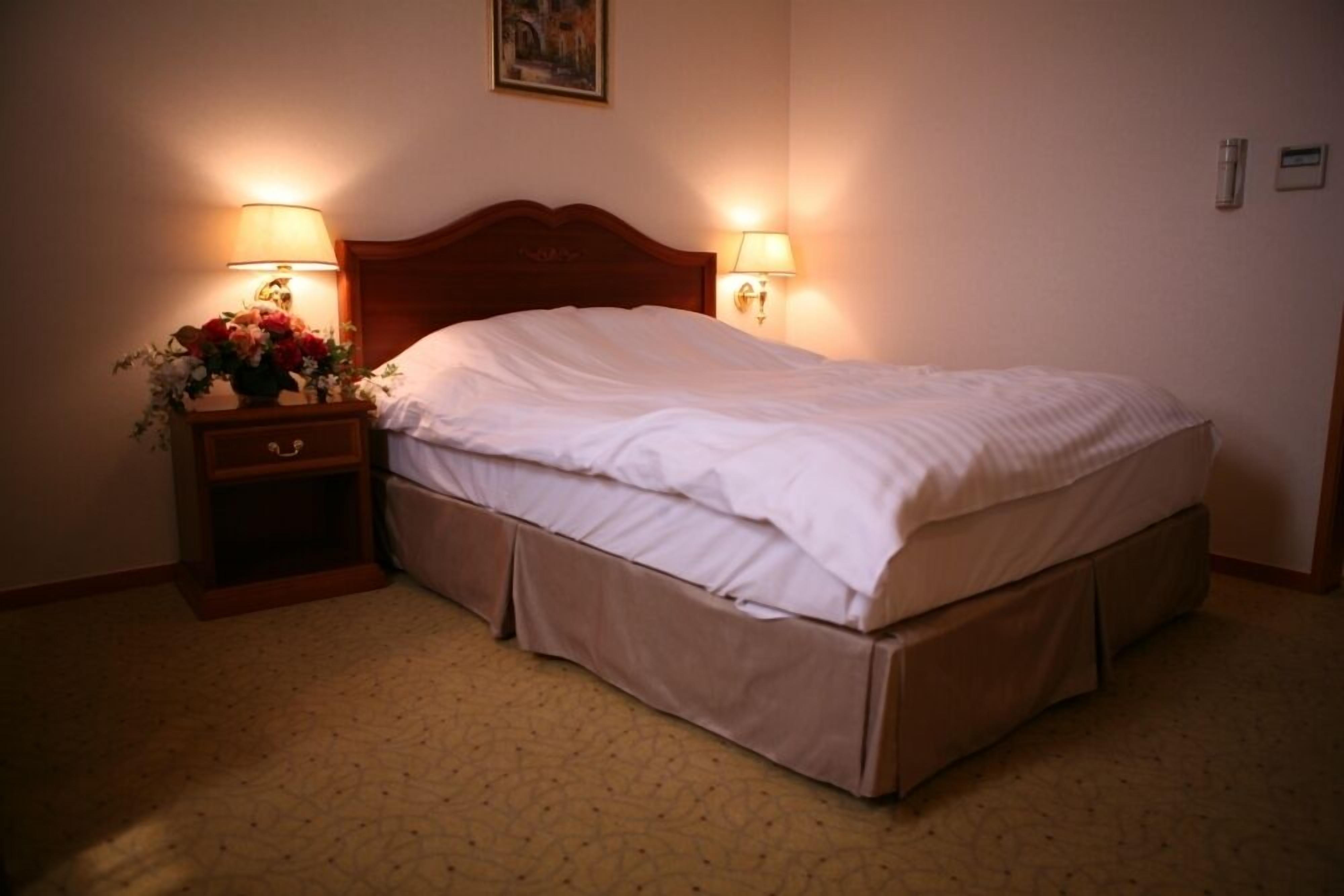 Habitación de hotel con una cama individual, dos lámparas de noche, una mesita de noche con un jarrón de flores y suelo cubierto de alfombra.