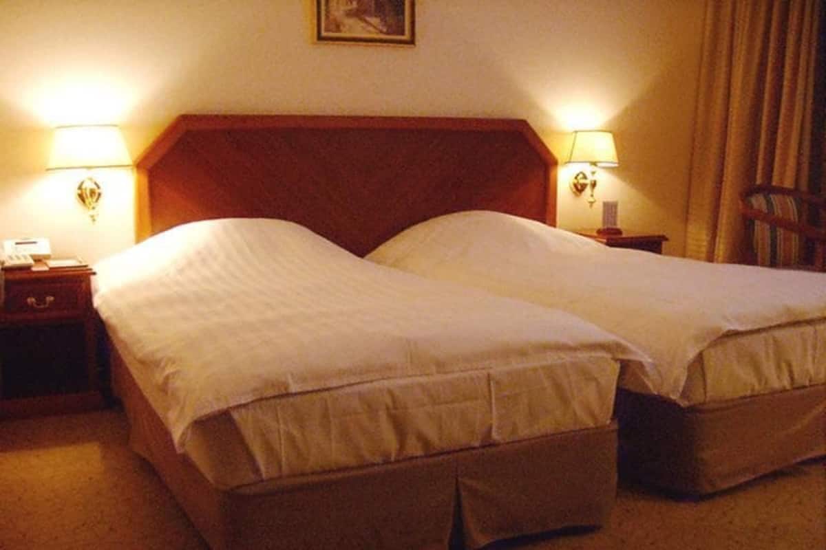 Habitación de hotel con dos camas individuales, cabecera de madera, lámparas de noche, mesitas de noche y una silla.