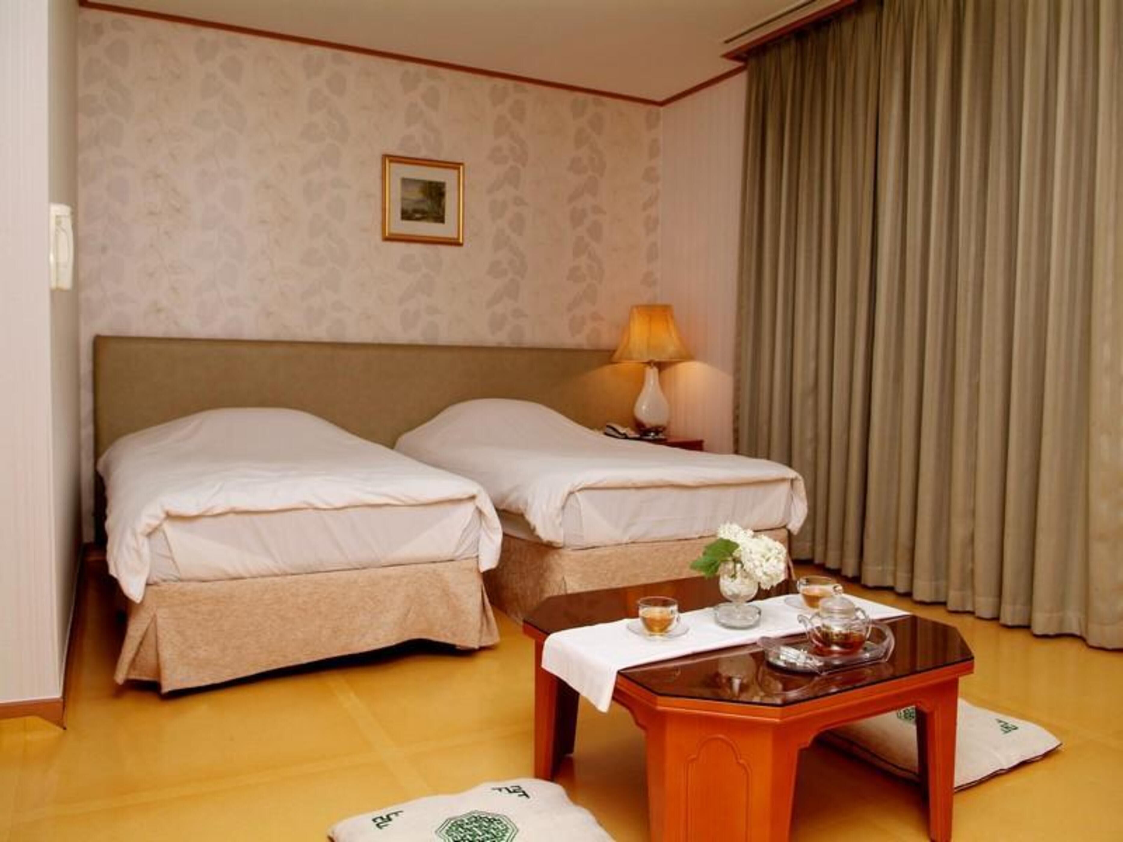 A hotel room with two beds, a nightstand with a lamp, a table with a teapot and cups, and a framed picture on the wall.