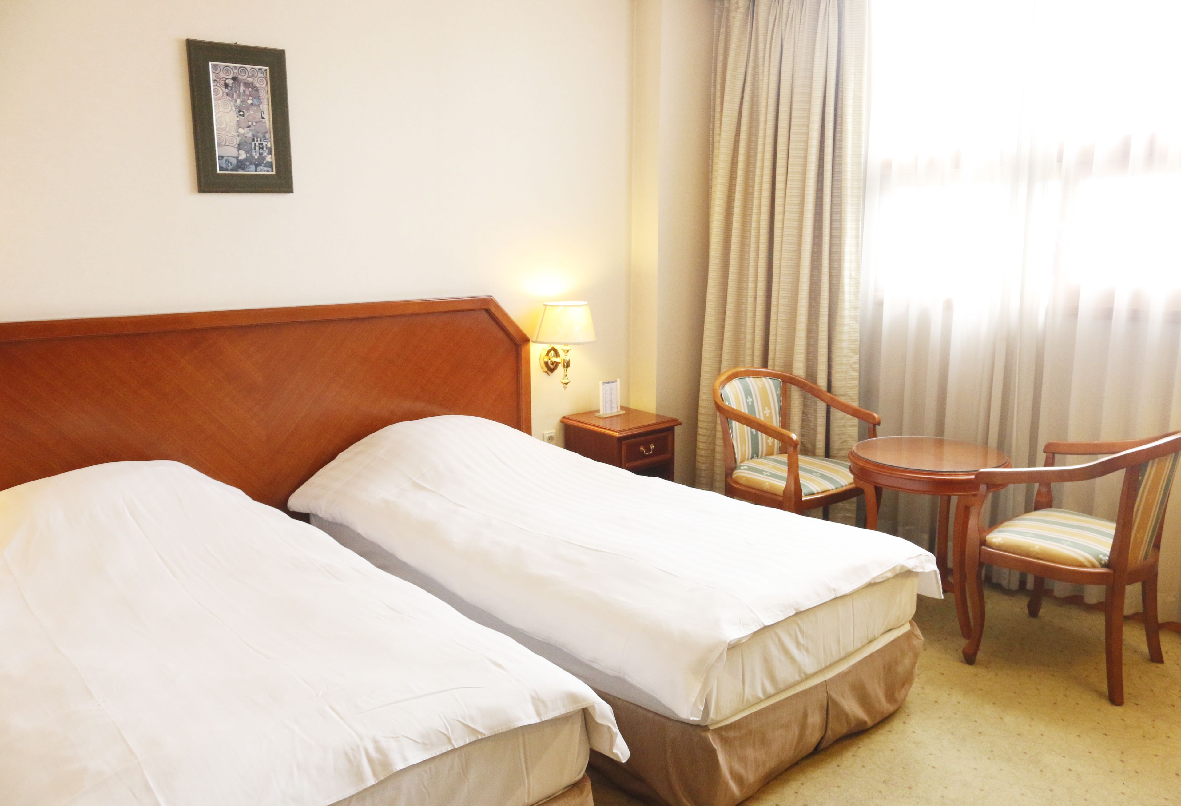 A hotel room with two beds, a wooden headboard, a nightstand with a lamp, a chair, a small table, and a window with curtains.
