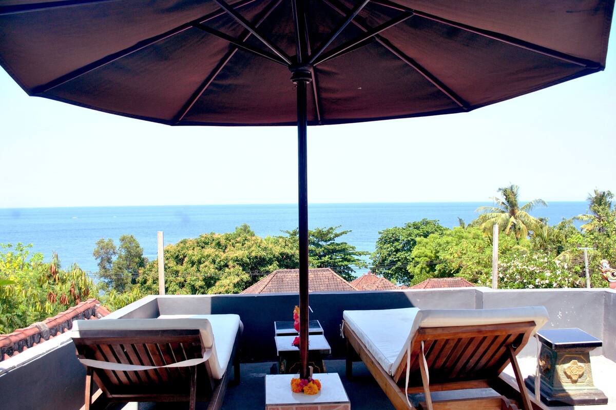 Un balcon avec vue sur l’océan, deux chaises en bois, une table et un grand parasol.