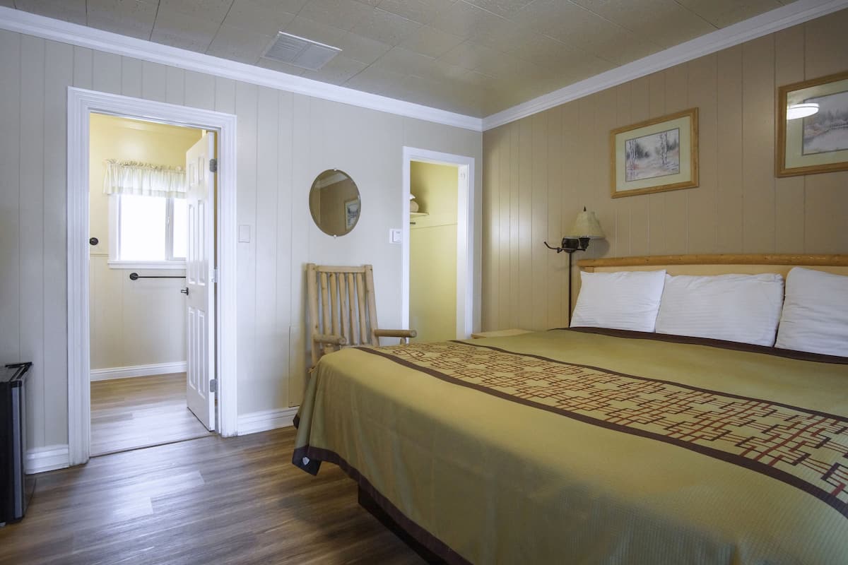 A room with a large bed, a chair, a door leading to another room, and a wall-mounted lamp.