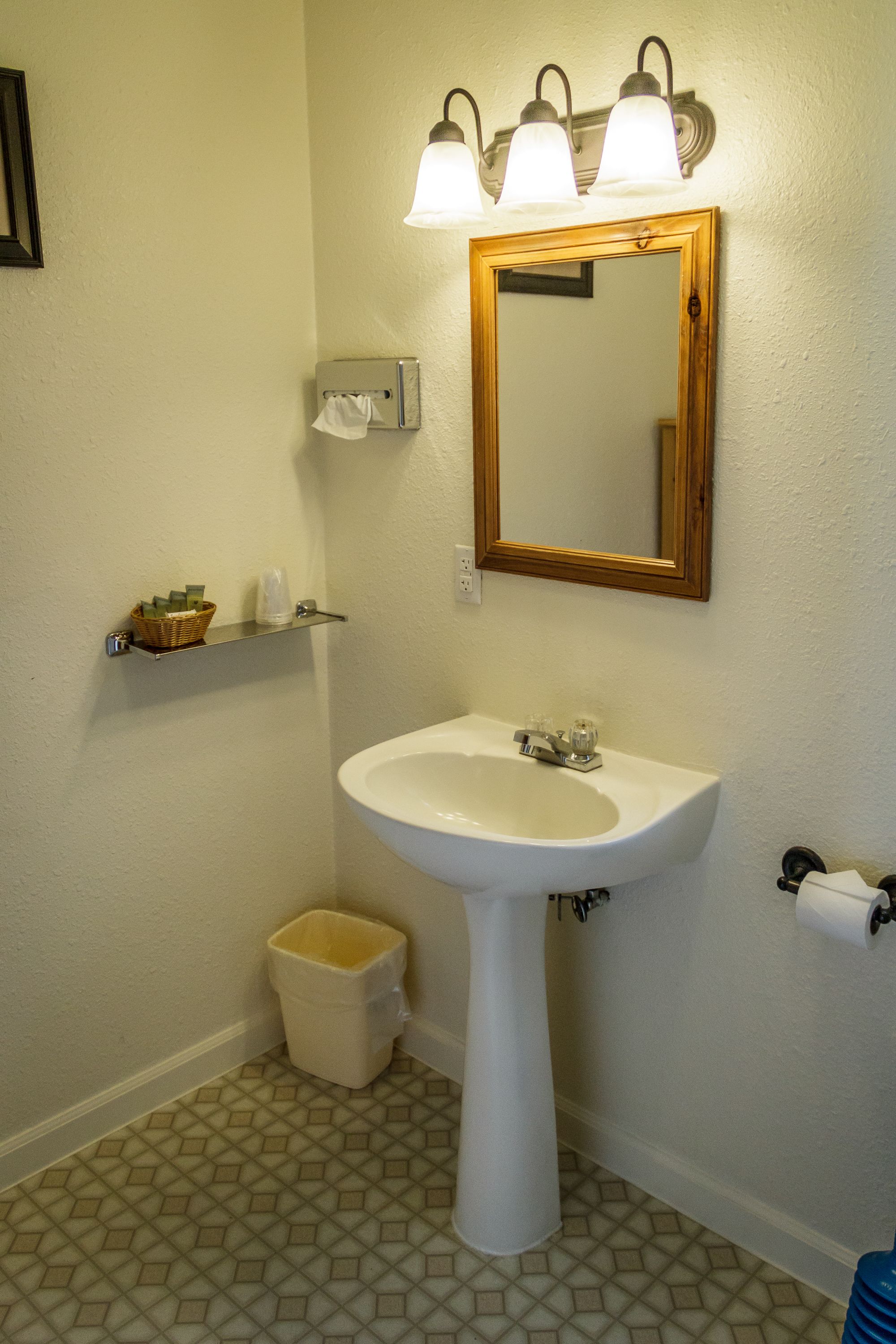 Un baño con lavamanos pedestal, tres luces empotradas, un espejo, un dispensador de papel higiénico, un cubo de basura y un estante con artículos de aseo.