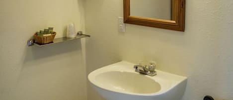 Un baño con lavamanos pedestal, tres luces empotradas, un espejo, un dispensador de papel higiénico, un cubo de basura y un estante con artículos de aseo.
